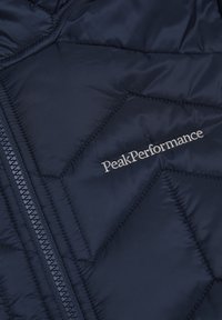 Námornícky puffer kabát s prešívaným dizajnom, obsahujúci viditeľný zips a vyšitý nápis "PeakPerformance" v striebornej farbe na oblasti hrudníka.