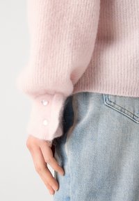 Light pink knitted sweater with a ribbed texture, featuring pearl buttons on the cuff, paired with faded blue denim jeans.