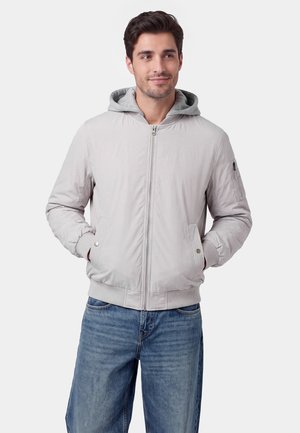 Mand med brunt hår iført en lysegrå lynlåstrøje med hættetrøje under og blå jeans, hænderne i lommerne.