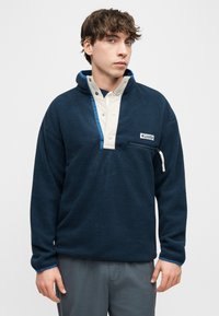 Νέος άνδρας που φοράει μπλε ναυτικό φλις με μπεζ κουμπωτό πατιλέτα και τσεπάκι στο στήθος με φερμουάρ, στέκεται μπροστά σε απλό φόντο.