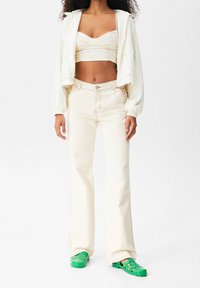 Kremowa krótka kurtka, fakturowany top, beżowe spodnie z wysokim stanem oraz zielone sandały, cechujące się casualowym i swobodnym stylem.