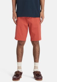 Pantaloni corti rossi in cotone con un taglio dritto, indossati sopra il ginocchio. Abbinati a calzini alla caviglia color crema e scarpe da barca marroni con lacci.