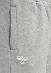 Zbliżenie na kieszeń z jasnoszarego materiału spodni z białym stylizowanym logo pszczoły w pobliżu szwu kieszeni.