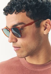 Jonge man met krullend donker haar, die schildpadkleurige zonnebril draagt en een gedempte rode trui met ronde hals, naar beneden kijkend tegen een witte achtergrond.