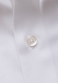 Valge riide lähivõte, millele on õmmeldud selge neljaauguline nööp lähedal kokkukeerdunud õmblusele.