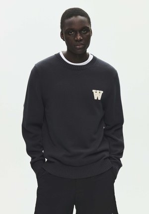 Ung mand iført en mørk crewneck-trøje med et hvidt "W"-logo på brystet, hænderne i lommerne, mod en enkel lys baggrund.