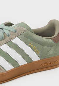 Zelené semišové tenisky Adidas Gazelle s bílými pruhy, béžovými akcenty, gumovou podrážkou a zlatým nápisem "Gazelle" na boku.