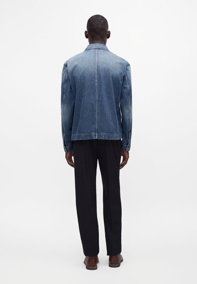 Homme debout, de dos à la caméra, portant une veste en jean bleue, un pantalon noir et des chaussures marron sur un fond clair uni.