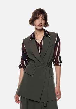 Donna con capelli castani corti indossa un blazer senza maniche belted di colore verde scuro sopra una camicia a righe bordeaux e crema con le maniche arrotolate, sfondo neutro.
