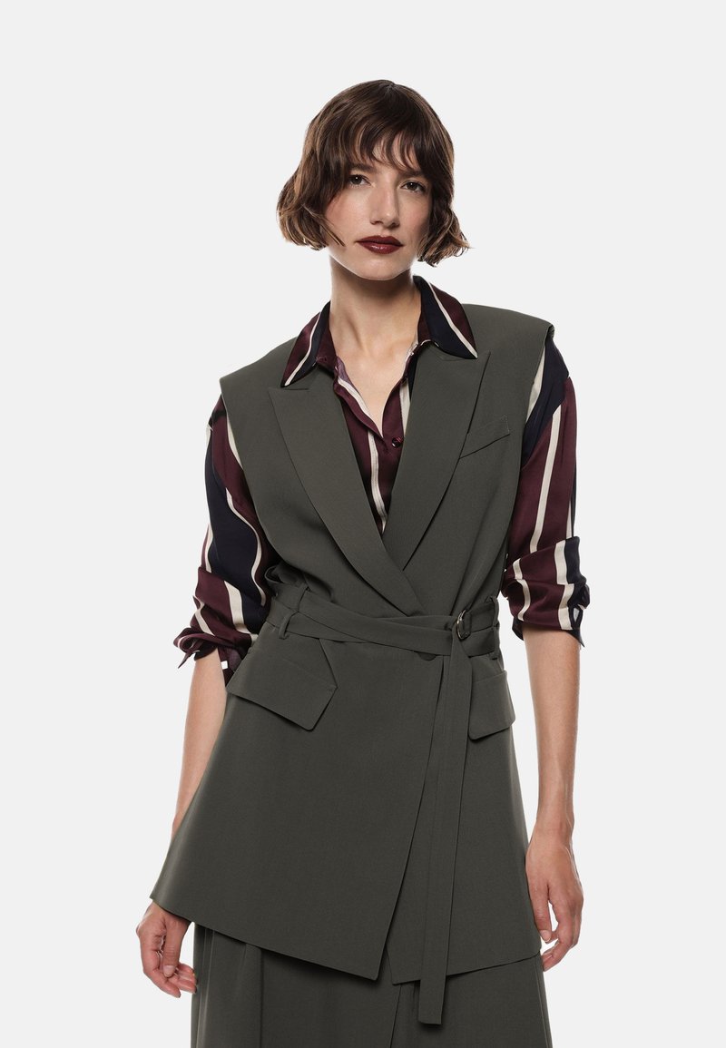Donna con capelli castani corti indossa un blazer senza maniche belted di colore verde scuro sopra una camicia a righe bordeaux e crema con le maniche arrotolate, sfondo neutro.