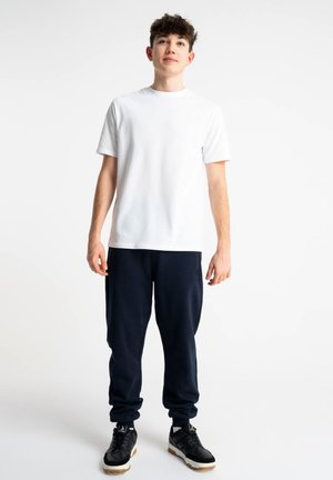 Młody mężczyzna stoi ubrany w prostą białą koszulkę, czarne dresy i czarne trampki na białym tle.