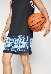 Basketball shorts with a blue geometric pattern, navy accents, and a Nike logo. Black sleeveless top. Basketball held in right hand.