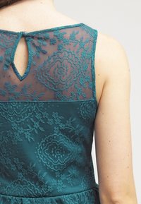 Tealfarbendes Spitzenkleid mit einem durchsichtigen Mesh-Oberteil, das mit Blumenmustern verziert ist, einem abgerundeten Rückenausschnitt und einem Knopfverschluss auf der Rückseite.
