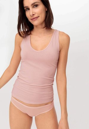Vrouw draagt een lichtroze mouwloos topje en bijpassend ondergoed, staand tegen een effen witte achtergrond, licht glimlachend.