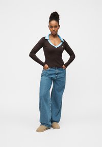 Γυναίκα που στέκεται με τα χέρια στις τσέπες, φοράει μαύρο μακρυμάνικο πουκάμισο με μπλε γιακά, τζιν με φαρδιά πόδια και μπεζ παπούτσια τύπου slip-on.