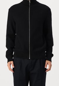 Pull zippé noir côtelé avec col haut, poignets ajustés et ourlet droit. Le tissu semble doux avec une finition texturée.