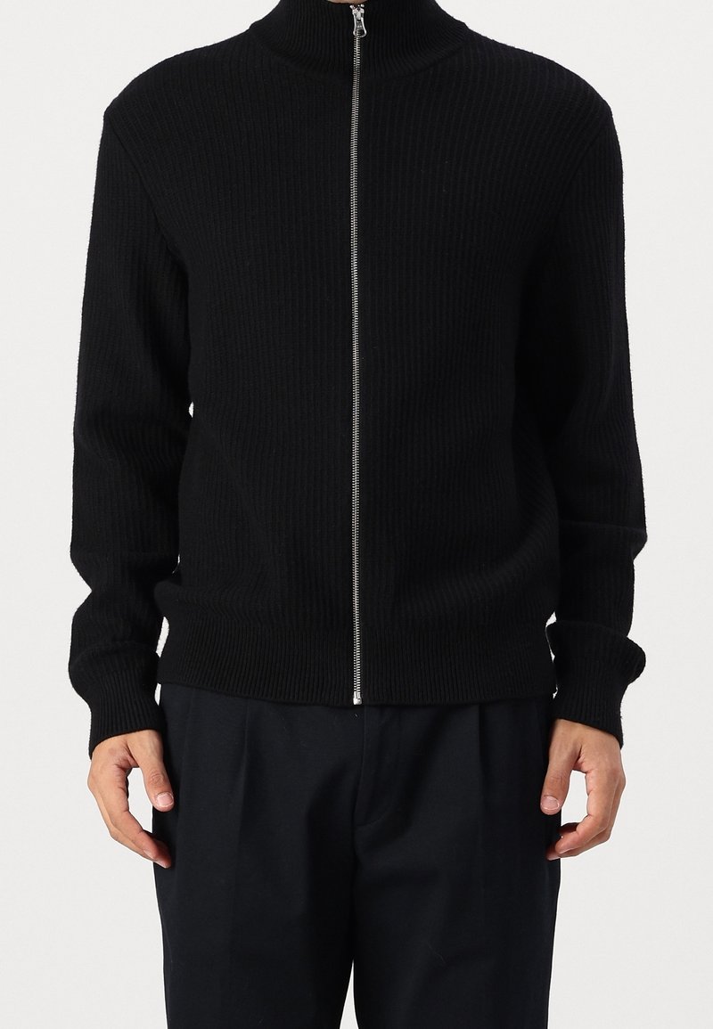 Pull zippé noir côtelé avec col haut, poignets ajustés et ourlet droit. Le tissu semble doux avec une finition texturée.