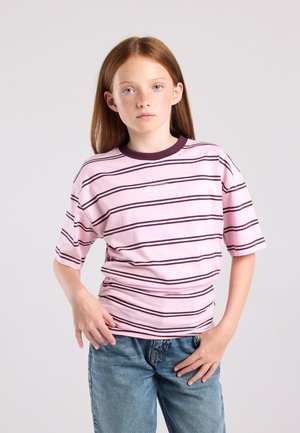 Jong meisje met rood haar, een roze en zwarte gestreepte trui en blauwe spijkerbroek, staat met de handen in de zij tegen een effen achtergrond.