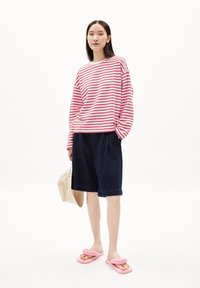 Rood-wit gestreepte langemouwshirt, donkere denimshorts met plooien en roze slipper. Model houdt een beige tas vast.
