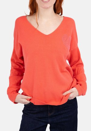 Pull orange corail avec un col en V et des manches longues, présentant un détail de cœur scintillant sur le côté gauche et des poignets côtelés.