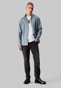 Camisa de botones a rayas azul claro, superpuesta sobre una camiseta gráfica blanca, combinada con jeans oscuros y zapatos negros, con un corte relajado.