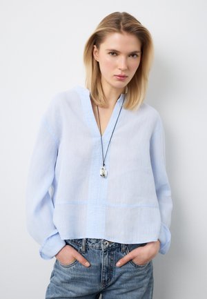 Frau trägt eine hellblaue, lockere Bluse, blaue Jeans und eine Anhänger-Halskette, steht mit den Händen in den Taschen vor einem weißen Hintergrund.