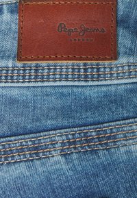 Niebieska tkanina dżinsowa z podwójnymi szwami i brązową skórzaną łatą z napisem "Pepe Jeans London" przyszytą do pasa.