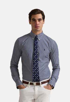 Camisa de dress a rayas azul marino y blanca con cuello abotonado, combinada con una corbata azul marino con patrones de escudo, vestida con pantalones blancos.