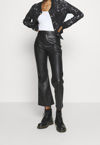 Musta pitsinen bomber-takki valkoisen tanktopin päällä, yhdistettynä korkeavyötäröisiin mustiin nahkahousuihin ja mustiin nauhakotuksiin.