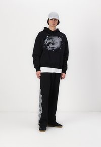 Felpa nera con grafica "Low Lights", abbinata a pantaloni della tuta neri con un design a fiamma argento. Il modello indossa un berretto grigio chiaro.