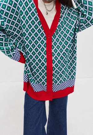 Person iført en grøn- og hvidternet cardigan med røde kanter og knapper, kombineret med blå jeans og et hjertevedhængs halskæde.