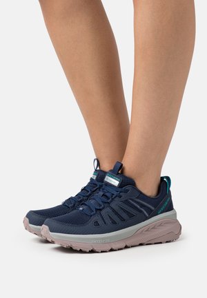 Jambes portant des baskets Skechers bleu marine à semelles beige clair, en maille respirante avec un design à lacets sur fond blanc.