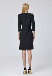 Robe noire structurée avec des manches trois quarts, taille ajustée et ourlet au genou. Portée avec des talons noirs brillants dotés d'accents dorés.