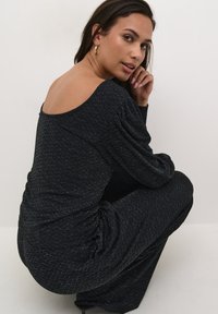 Schwarzer Jumpsuit mit fließendem Design, ausgestattet mit einem tiefen Rückenausschnitt, langen Ärmeln und einem strukturierten Stoff, der durchgehend metallisch goldene Fäden aufweist.