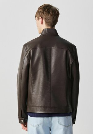 Junger Mann trägt eine dunkelbraune Lederjacke und hellblaue Jeans, steht mit dem Rücken zur Kamera vor einem schlichten hellgrauen Hintergrund.