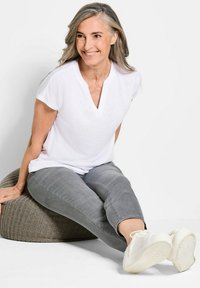 T-shirt bianca a scollo a V, maniche corte, abbinata a jeans grigi, seduta su un cuscino tondo grigio con texture, indossando sneakers bianche.