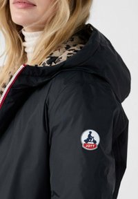Zwarte lichtgewicht jas met een patroon aan de binnenkant. Heeft een rode rits en een rond logo op de mouw. Het materiaal lijkt glad en waterafstotend te zijn.