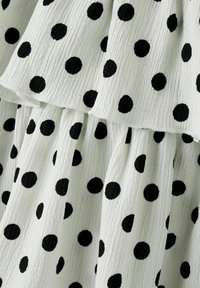 Vit texturerad tyg med svarta prickar som har en samlad, lager-på-lager konstruktion som skapar mjuka veck och volym.