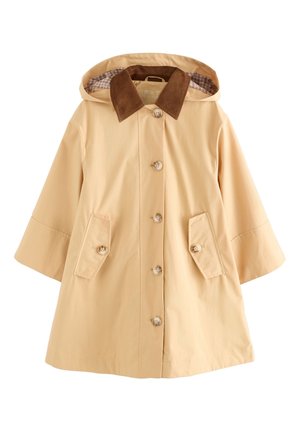 Unge iført en beige frakke med knapper og brun krave samt blå jeans, stående mod en ensfarvet lys baggrund.
