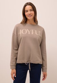 Kvinna iförd en taupe sweatshirt med texten "JOYFUL" och snörning i nederkant, kombinerat med mörkblå jeans, stående mot en enfärgad beige bakgrund.