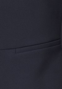 Tissu bleu marine avec une texture lisse et un léger éclat. Comprend un détail de poche verticale fine unique sur le côté droit.