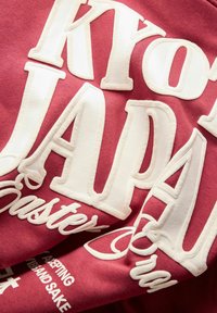 Bordeauxrode sweatshirt met grote witte, gestructureerde letters "KYOTO JAPAN" en kleinere tekst eronder, gemaakt van zacht katoen en met een casual ontwerp.