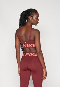 Bordo športni modrček s tankimi naramnicami, opremljen z belim logotipom Nike, skupaj z ujemajočimi se visokimi hlačami z izrazitim oranžnim napisom. Gladka tekstura.