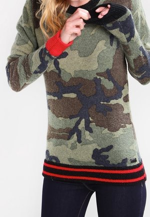 Persona con un suéter de patrón camuflaje con puños y dobladillo a rayas rojas y negras, ajustándose el escote con ambas manos.