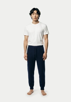 Junger Mann steht barfuß, trägt ein weißes T-Shirt mit kurzen Ärmeln und marineblaue gestrickte Hose mit Kordelzug vor schlichtem Hintergrund.