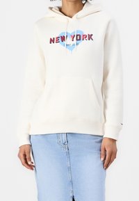 Mujer llevando una sudadera color crema con la palabra "New York" y un gráfico de corazón azul, combinada con una falda de mezclilla azul claro.
