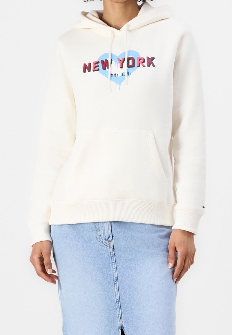 Mujer llevando una sudadera color crema con la palabra "New York" y un gráfico de corazón azul, combinada con una falda de mezclilla azul claro.