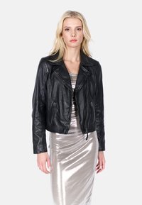 Zwart leren bikerjack met ritsen, cropped ontwerp, pieklapels en textuurafwerking. Draagt over een metallic zilveren jurk.