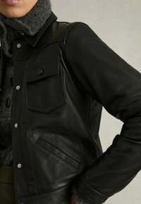 Schwarze Lederjacke mit Druckknöpfen, grauem Strickkragen, zwei Fronttaschen mit Klappen und glatter Textur. Detaillierte Nähte an den Taschen.