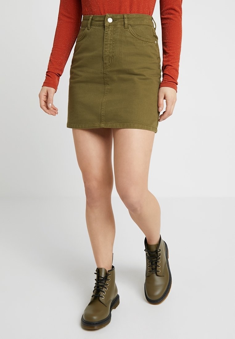 Falda de mezclilla verde olivo con bolsillos frontales y lazos para cinturón, combinada con botas de tobillo negras con cordones. Textura suave y diseño ajustado.
